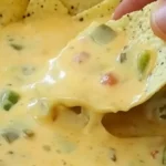 A tortilla chip dipped into The Greatest Queso That Ever Lived, showing creamy texture with melted cheese and diced jalapeños.